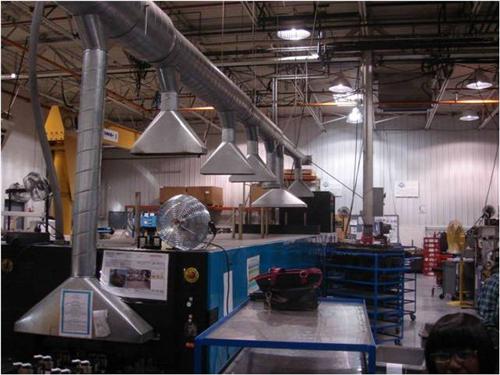 Industrial shop floor with overhead ductwork and funnel-shaped extraction hoods above workstations, with fans and storage racks visible.